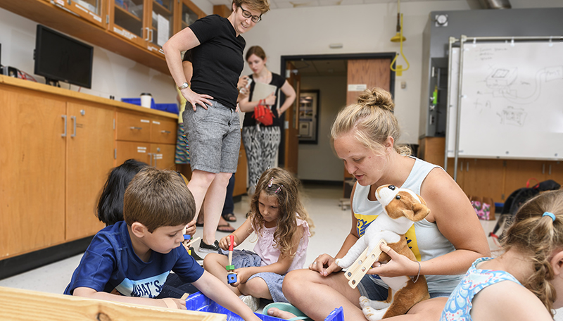 The Lab School's Body Mechanics camp (ages 4-7) are going into Jenny Buckley's engineering lab and learning about the skeleton, working with xrays, 'fixing' broken bones, etc., the students will design prosthetics for a stuffed dog. ID's: UD students Grey top, hair in bun – Makenzie Mullen ECE major Grey top long brown hair – Jordyn Schrader – bio engineering White/pink peasant blouse – Danielle Gerstman – mech engineering alum Blue UD heart shirt – Erin Rezich – mech engineering LPS camp Kids Boys Blue Tshirt (fixed dog’s leg) Sean White shirt orange shorts – Alex Green plaid shirt – Aaryan Girls White shirt – Mehr Pink shirt Ella Blue patterned shirt Emerson Blue shirt purple shorts – Izzie (Who may have limited permission, so we have to show to LPS.)