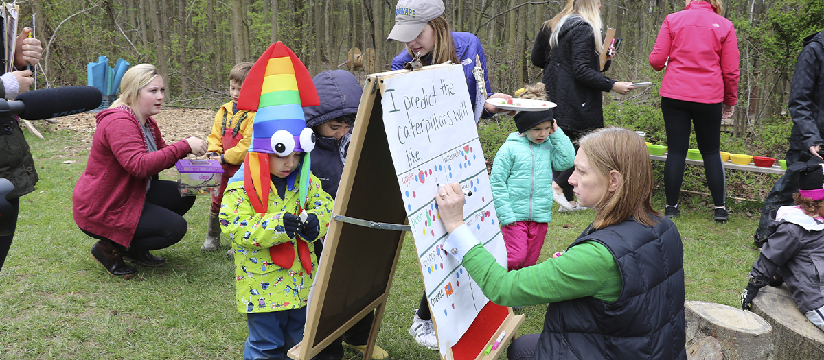 Professor Jen Gallo Fox and Lab School teacher help student do caterpillar activity outside